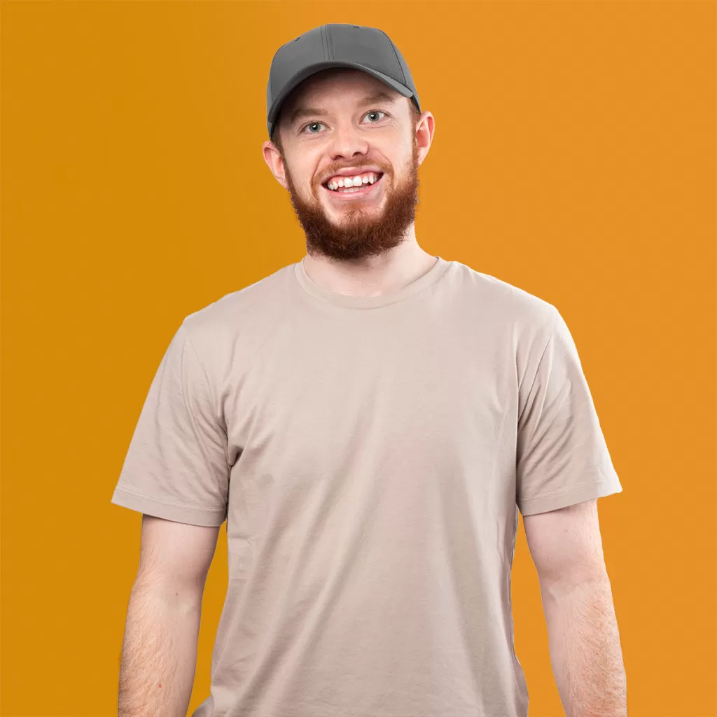 Man with red hair and beard, wearing green cap and beige tshirt faces the camera and smiles