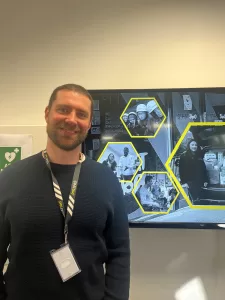 Man with brown hair and beard, wearing black long sleeved jumper and lanyard, standing in front of a TV screen faces the camera and smiles.